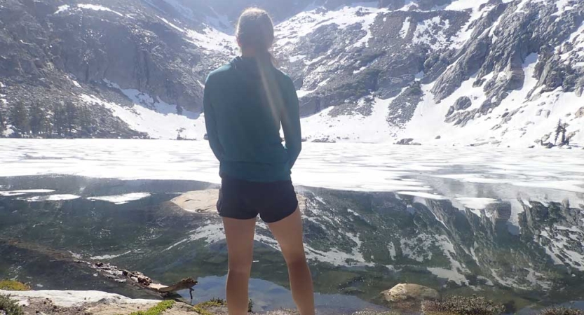 A person faces away from the camera and looks out at an alpine lake, reflecting the snow capped mountains surrounding it. 